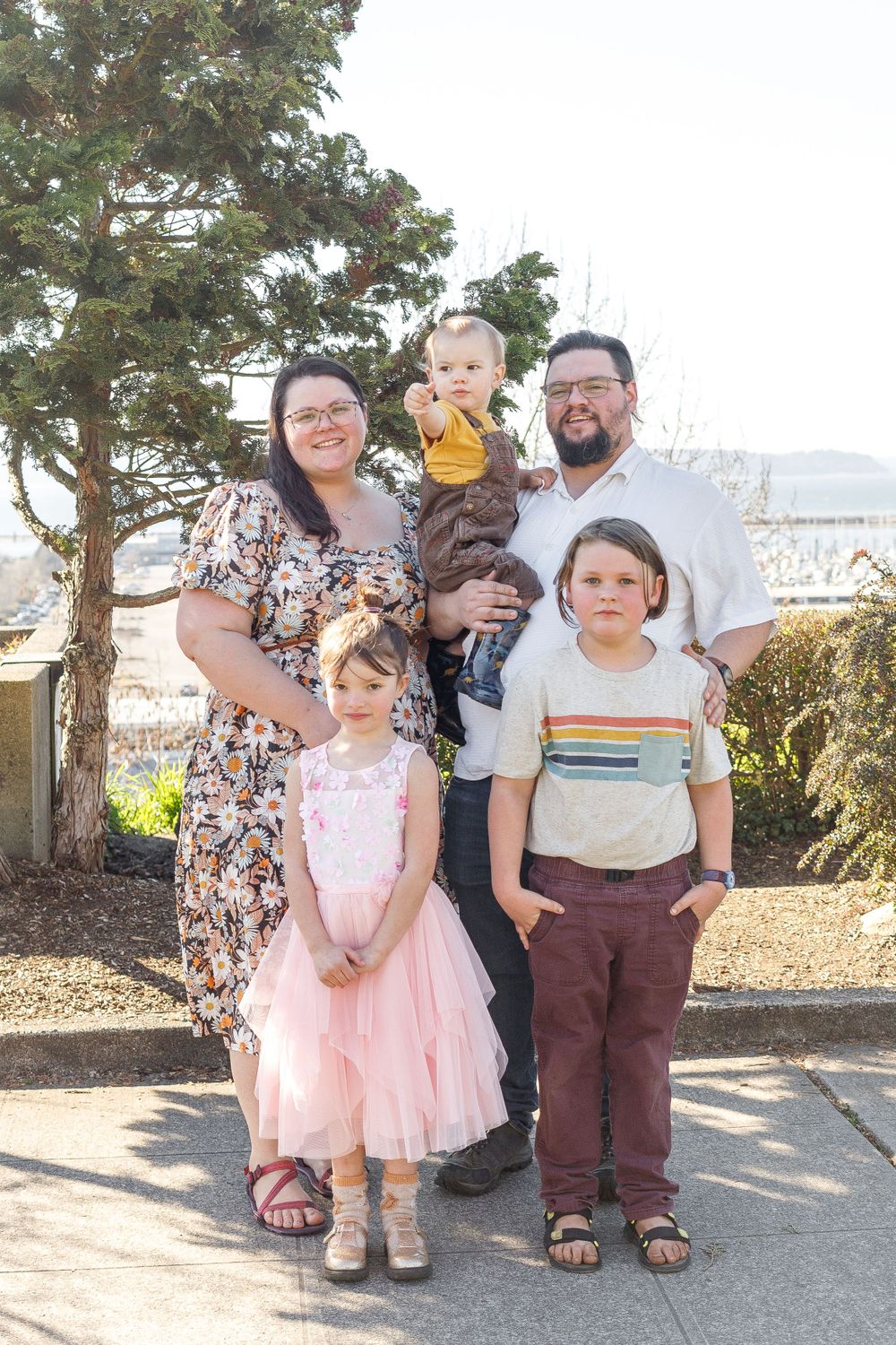 A family of five poses together on a sunny day in front of evergreen trees.