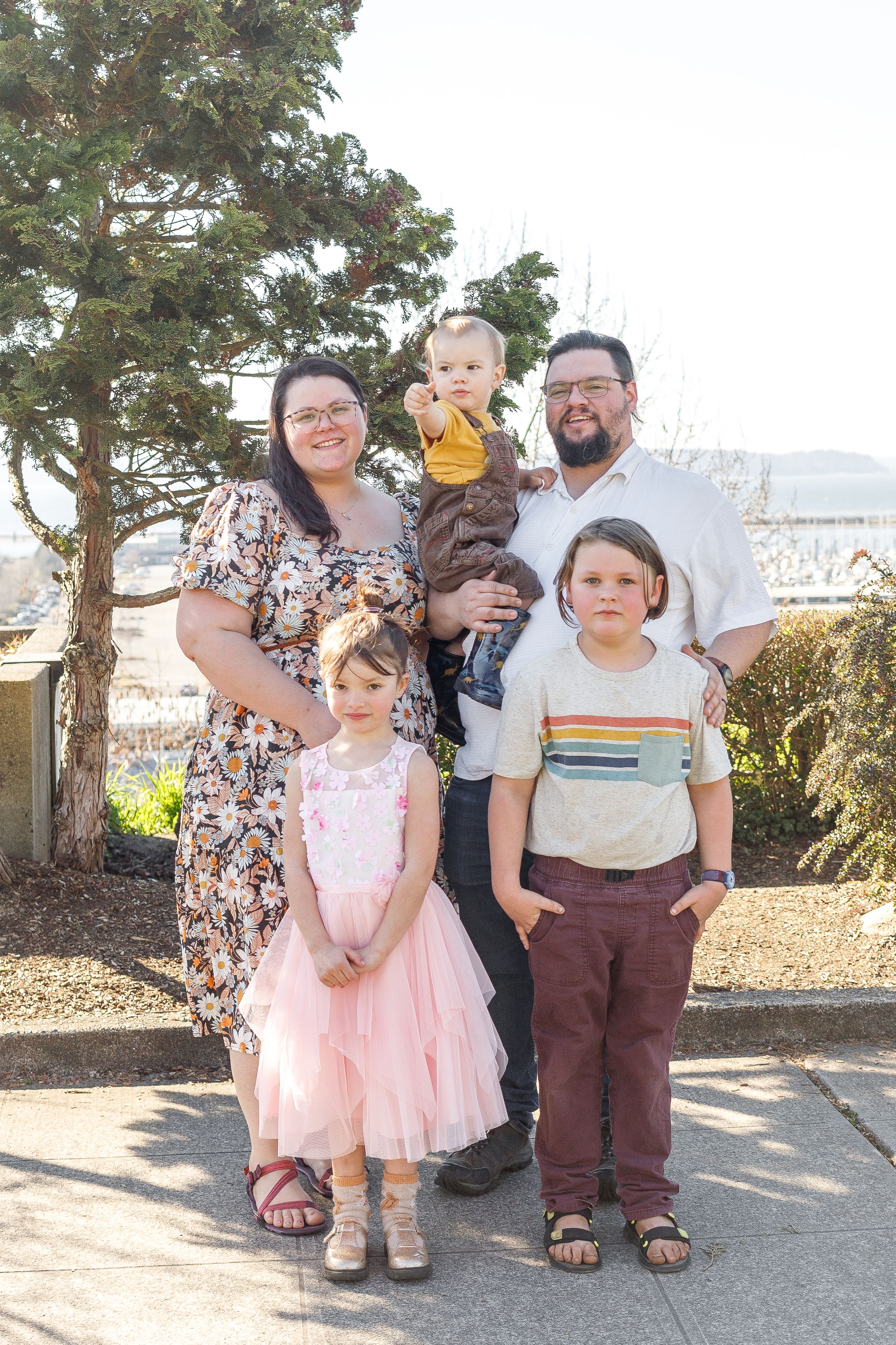 A family of five poses together on a sunny day in front of evergreen trees.