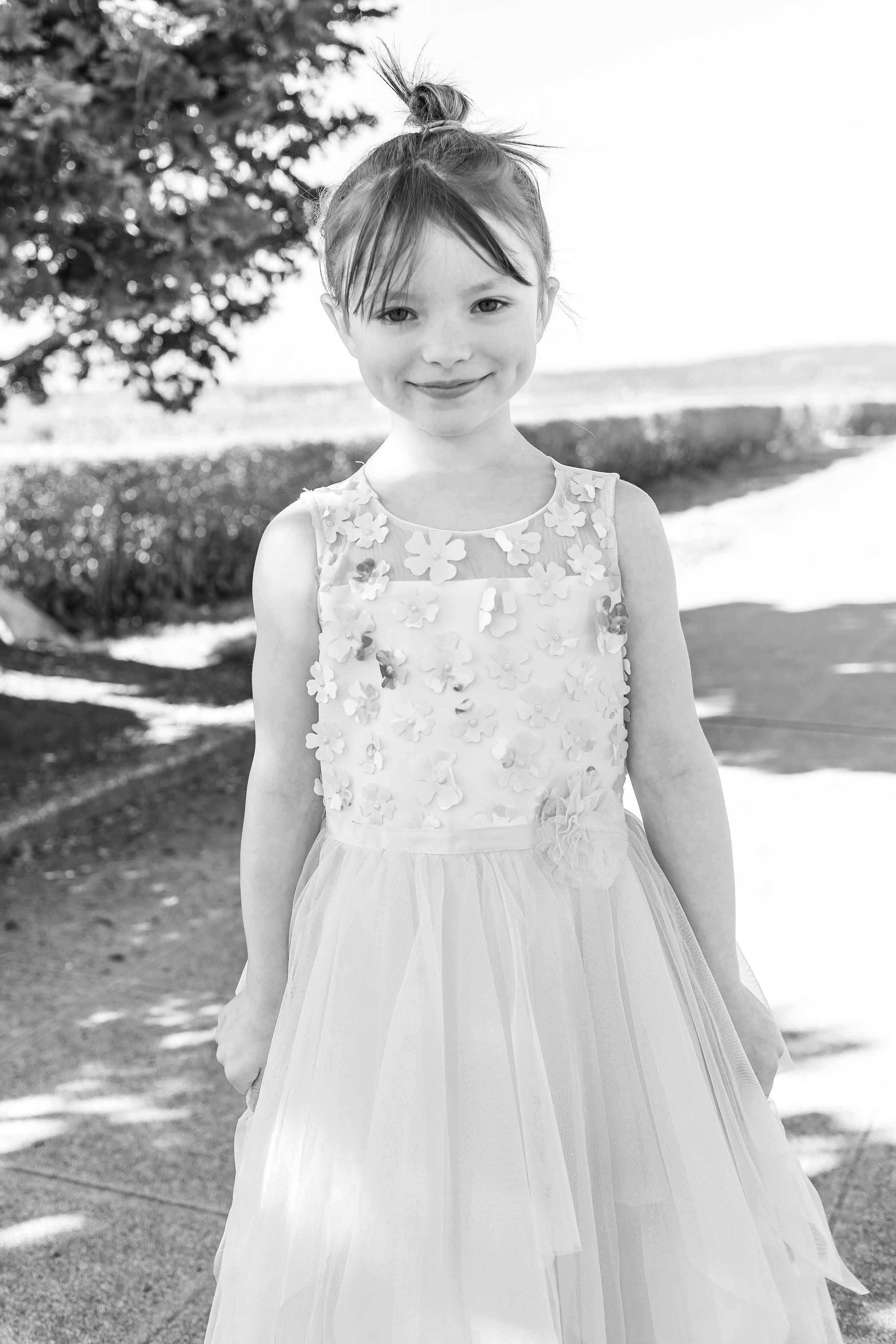 A young child in a light colored dress stands outside on a sunny day.