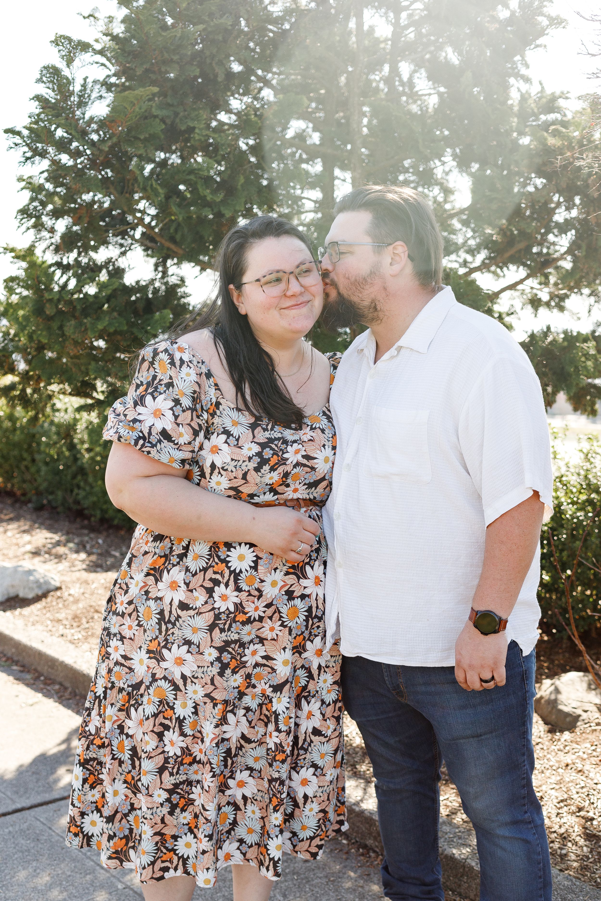 A couple shares a tender moment outdoors in front of greenery on a sunny day.