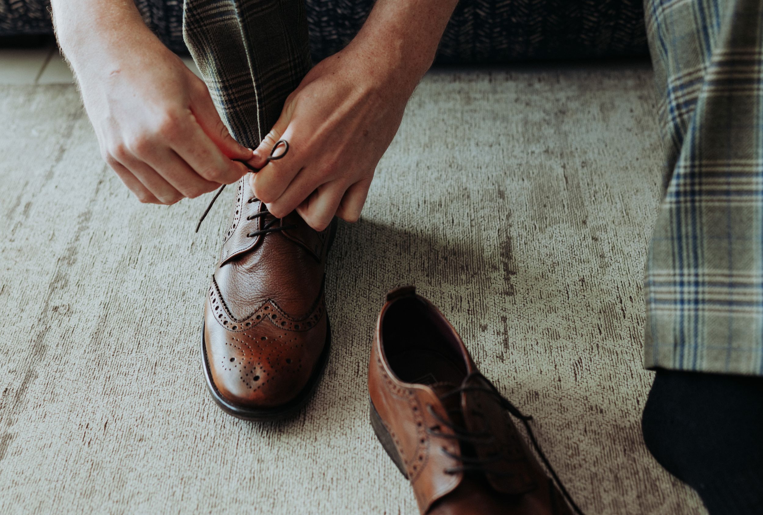 Hands tying laces on brown leather brogue dress shoes on a gray floor.