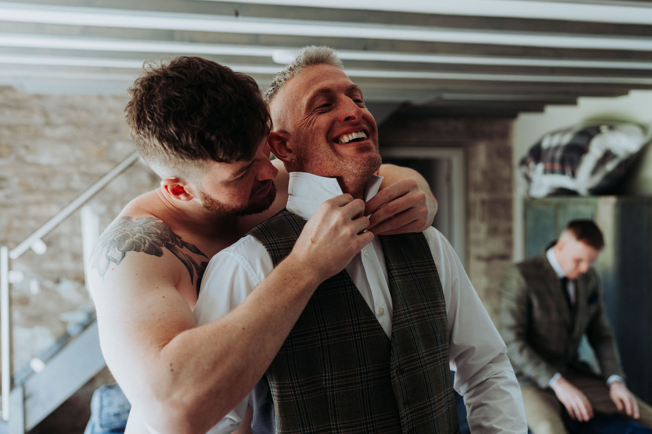 Two people adjusting formal attire and laughing together during wedding preparations.