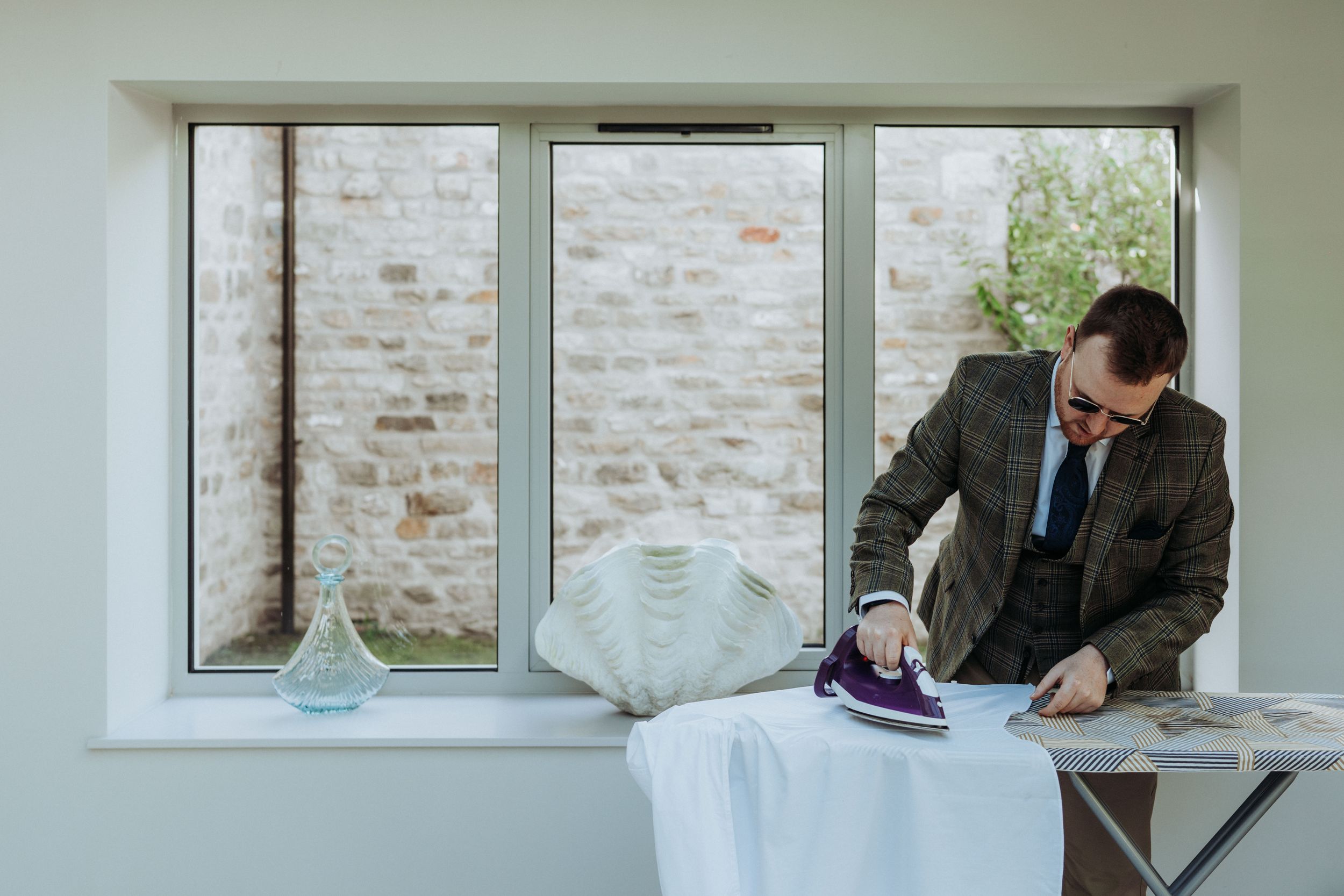 Person in suit jacket ironing white dress shirt near window with exposed brick wall.
