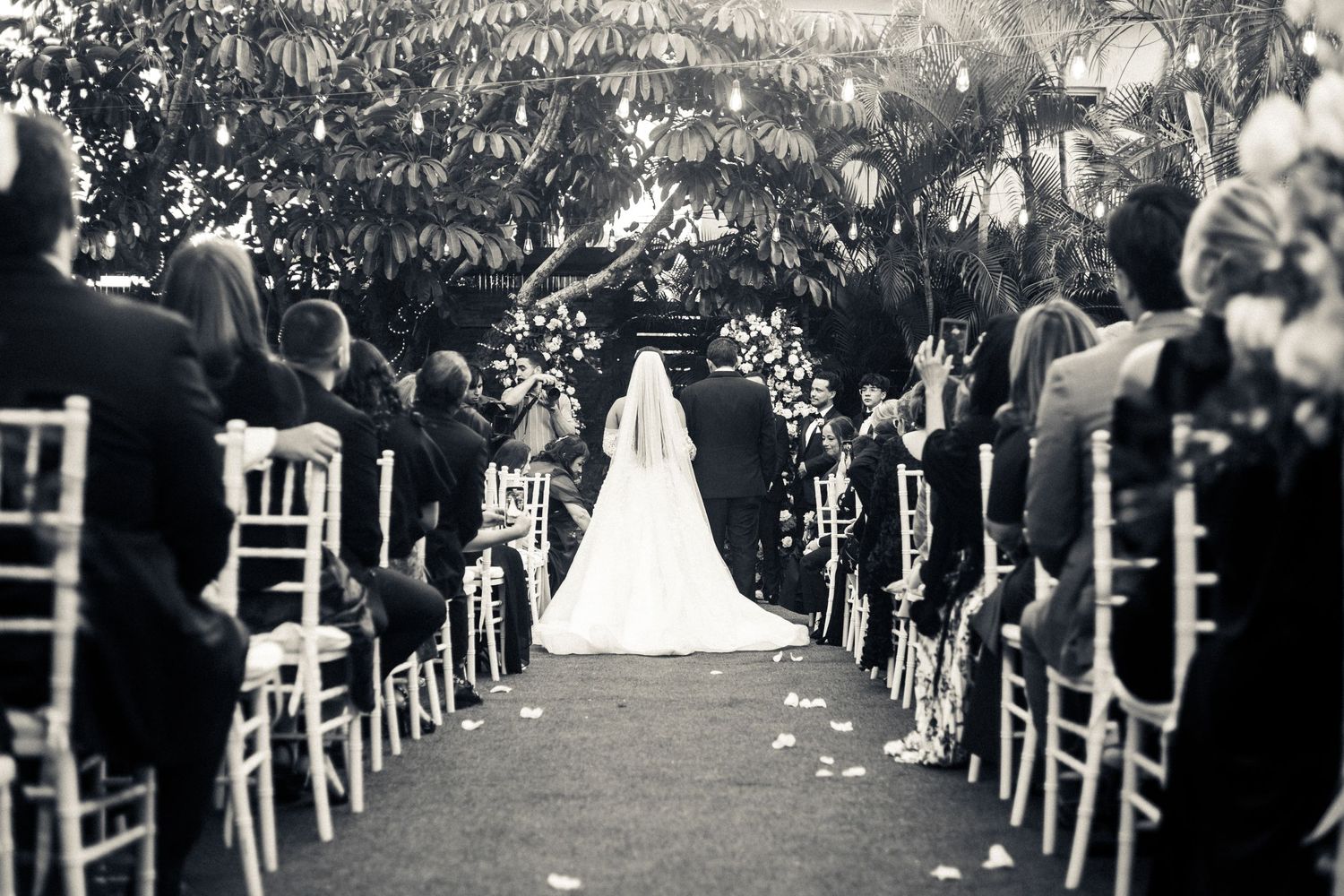 Black and white photo of a wedding ceremony with guests seated along a floral-lined aisle.
