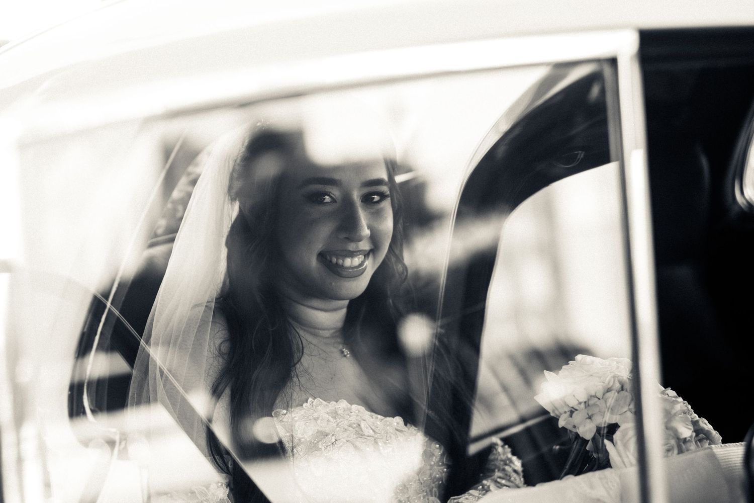 A black and white photograph of a bride smiling through a car window.