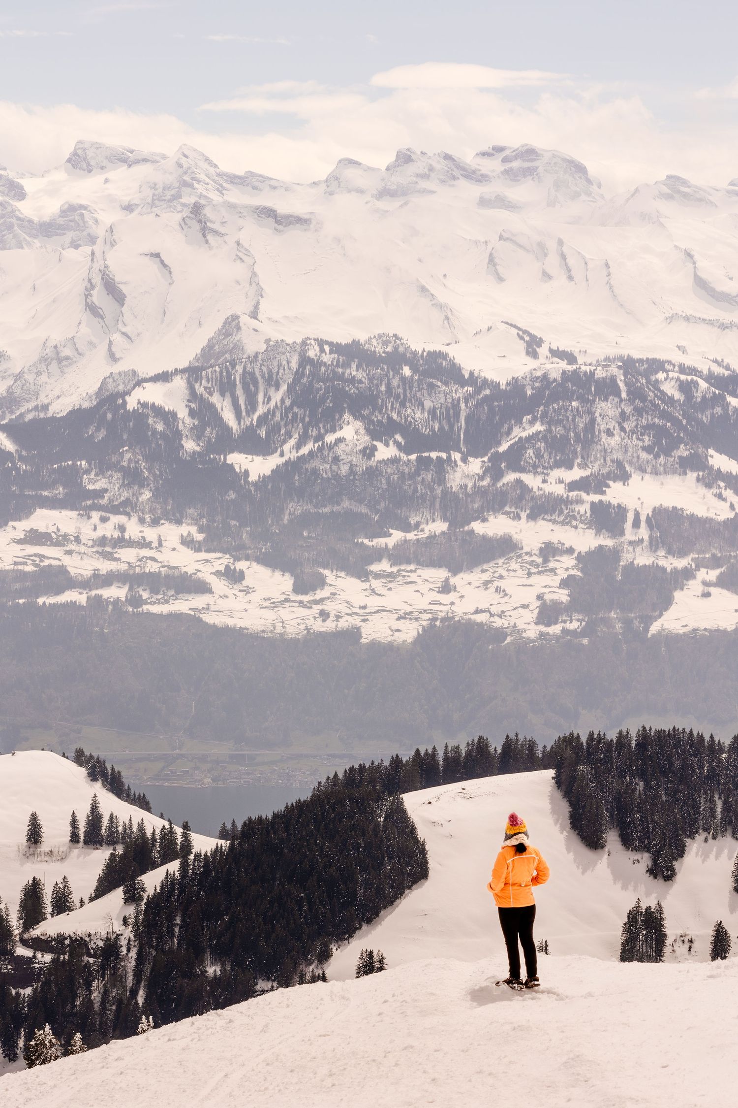 A skier in bright orange jacket stands on snowy mountain peak overlooking dramatic Alpine landscape with snow-capped peaks.