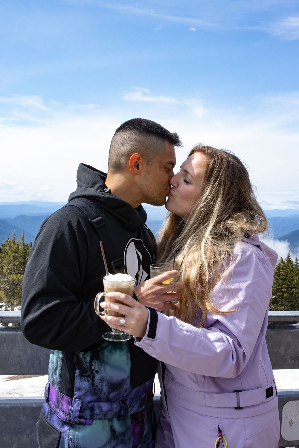 Couple kissing at Timberline Lodge while holding drinks, with scenic blue sky and mountain range in the background.