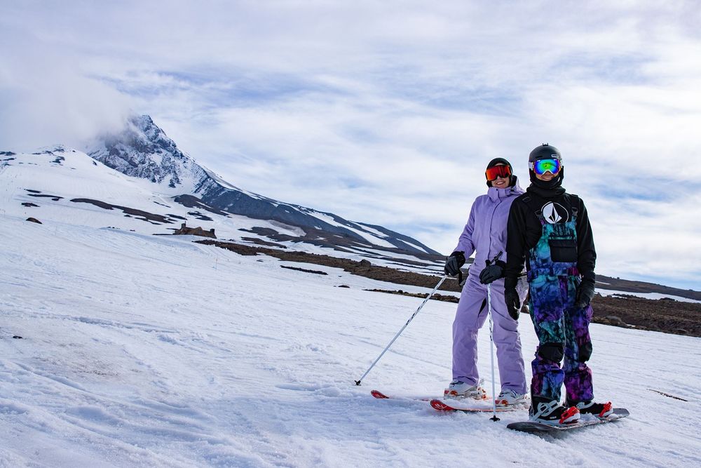 Two skiers pose on a snowy mountain slope with a dramatic peak and cloudy sky in the background.
