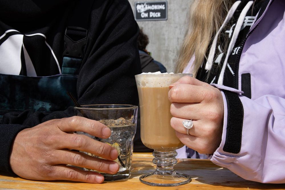 Two people sitting at Timberline Lodge holding coffee drinks, one wearing an engagement ring, with a small glass nearby.