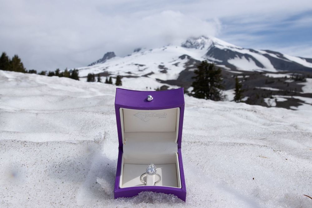 Diamond engagement ring in purple box displayed in snow with snowy Mt.Hood and evergreen trees in background.