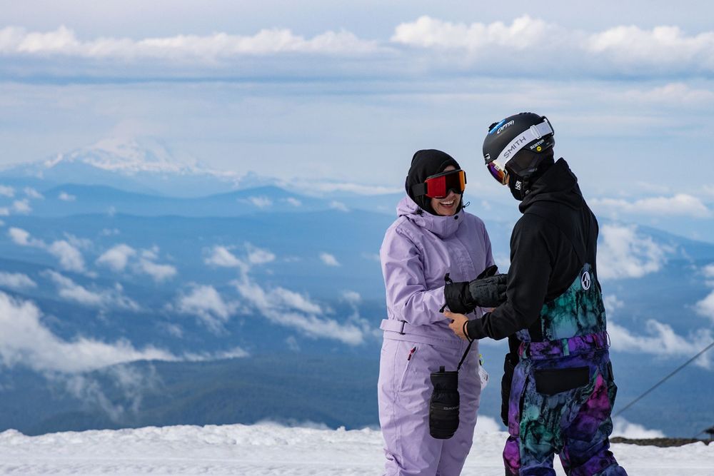 Two snowboarders in colorful winter gear hold hands on a snowy mountain summit with clouds and peaks below.