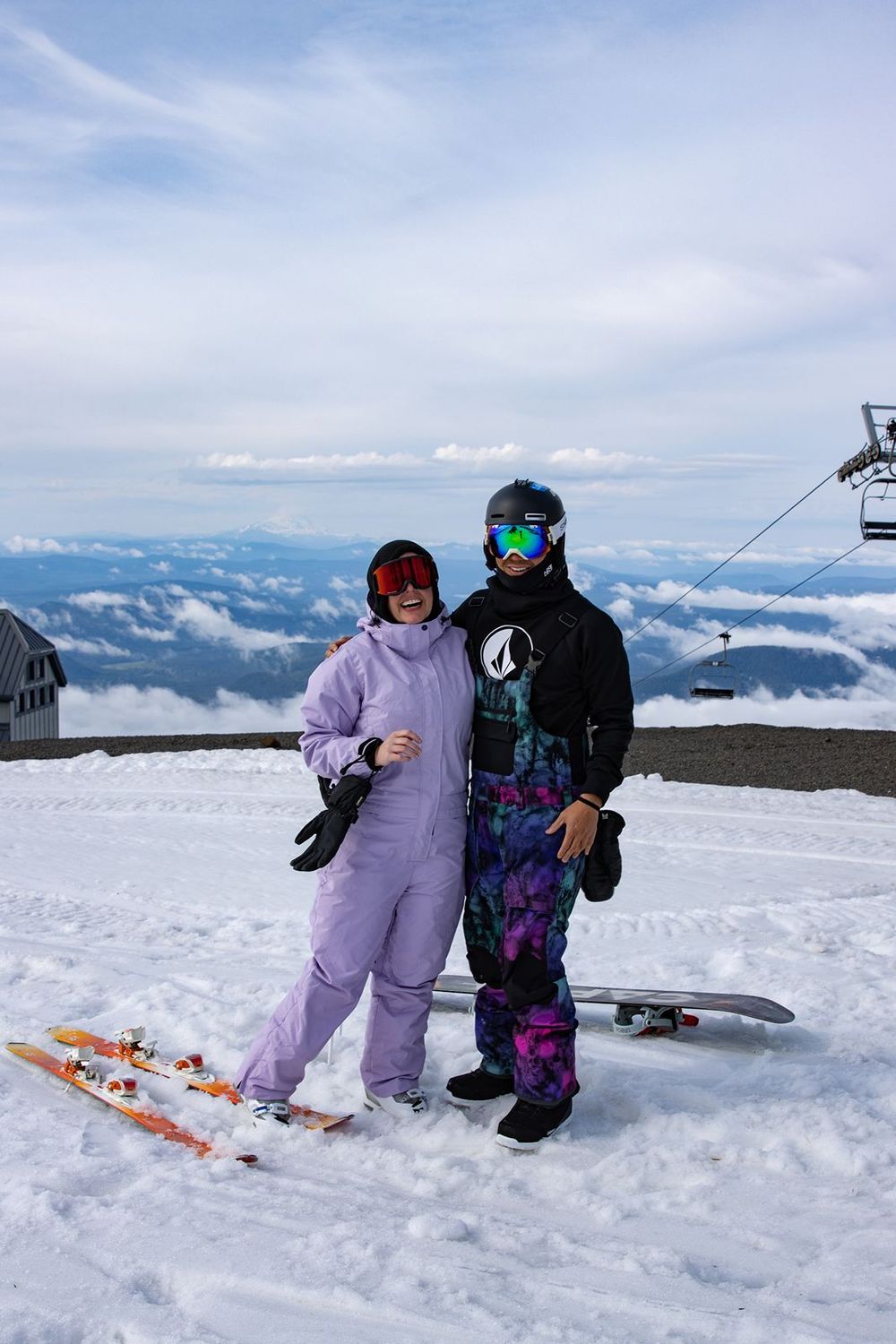 Two snowboarders in colorful gear pose on a snowy mountain summit above the clouds on a sunny day.