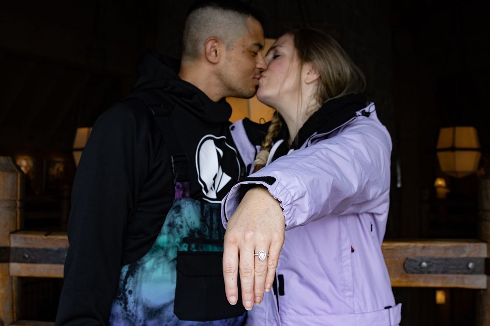 Couple sharing a kiss while woman displays engagement ring, wearing casual jackets in Historic Timberline Lodge.