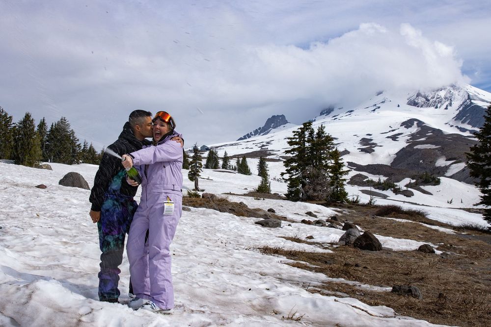 Couple embracing on a snowy Mount Hood slope with evergreen trees and cloudy peaks in the background.