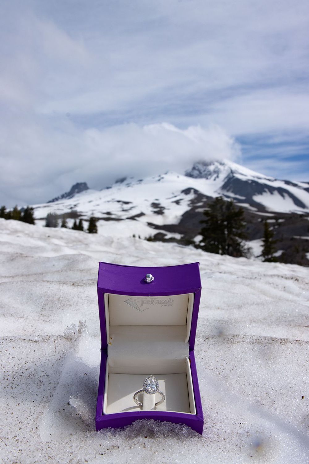 Purple ring box with diamond engagement ring sitting in snow with snowy Mount Hood backdrop.
