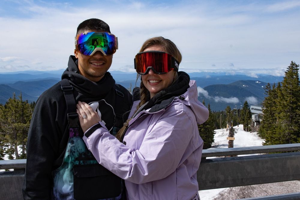 Two skiers in colorful goggles and winter gear pose smiling at a snowy Timberline Lodge with scenic views.