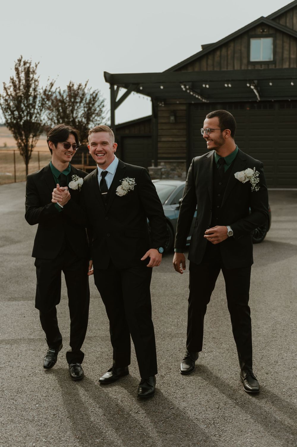 Groomsmen in black suits sharing a laugh outside rustic venue.