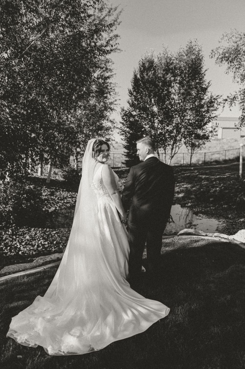 Classic black and white wedding photo of couple in elegant attire with flowing veil against garden backdrop.