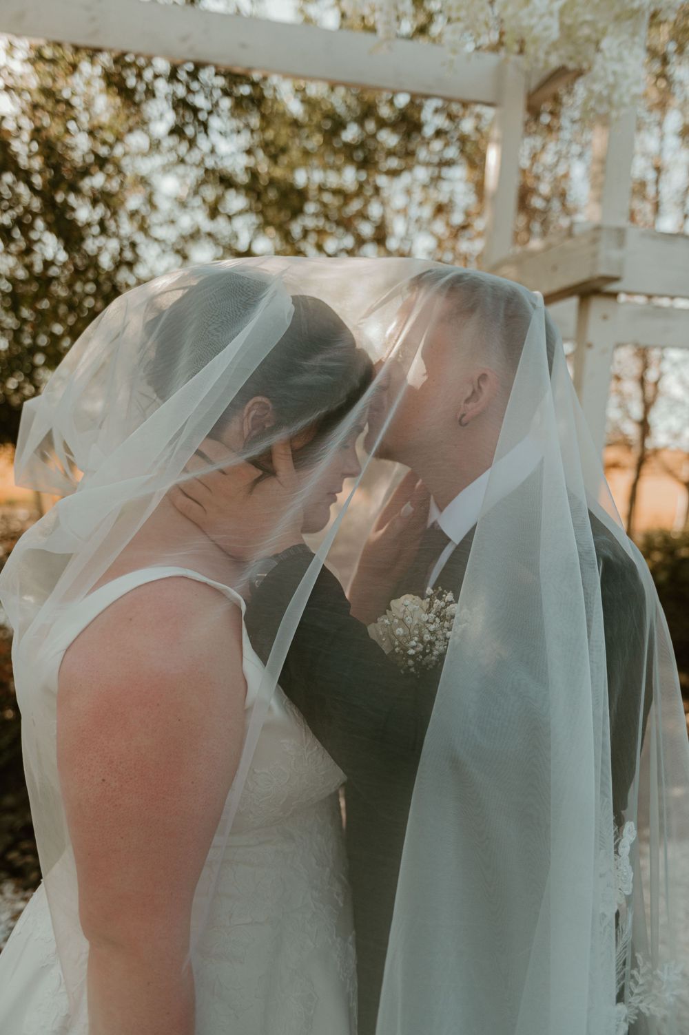 Intimate wedding moment of two brides sharing a kiss under veils at sunset.