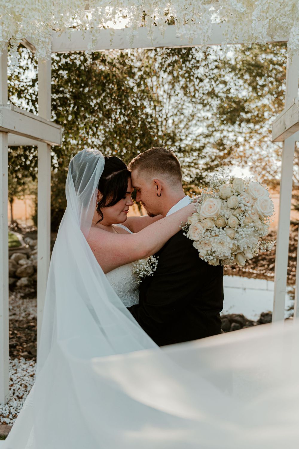 Romantic wedding portrait of couple embracing under white gazebo surrounded by flowers.