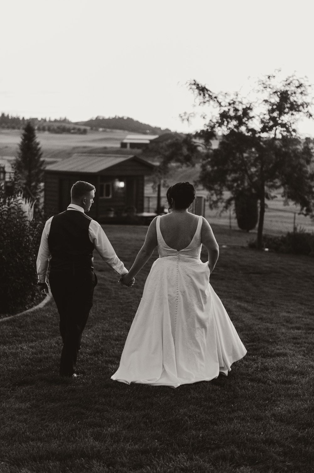 Newlyweds walking hand in hand through a grassy field at dusk with rustic buildings in the background.
