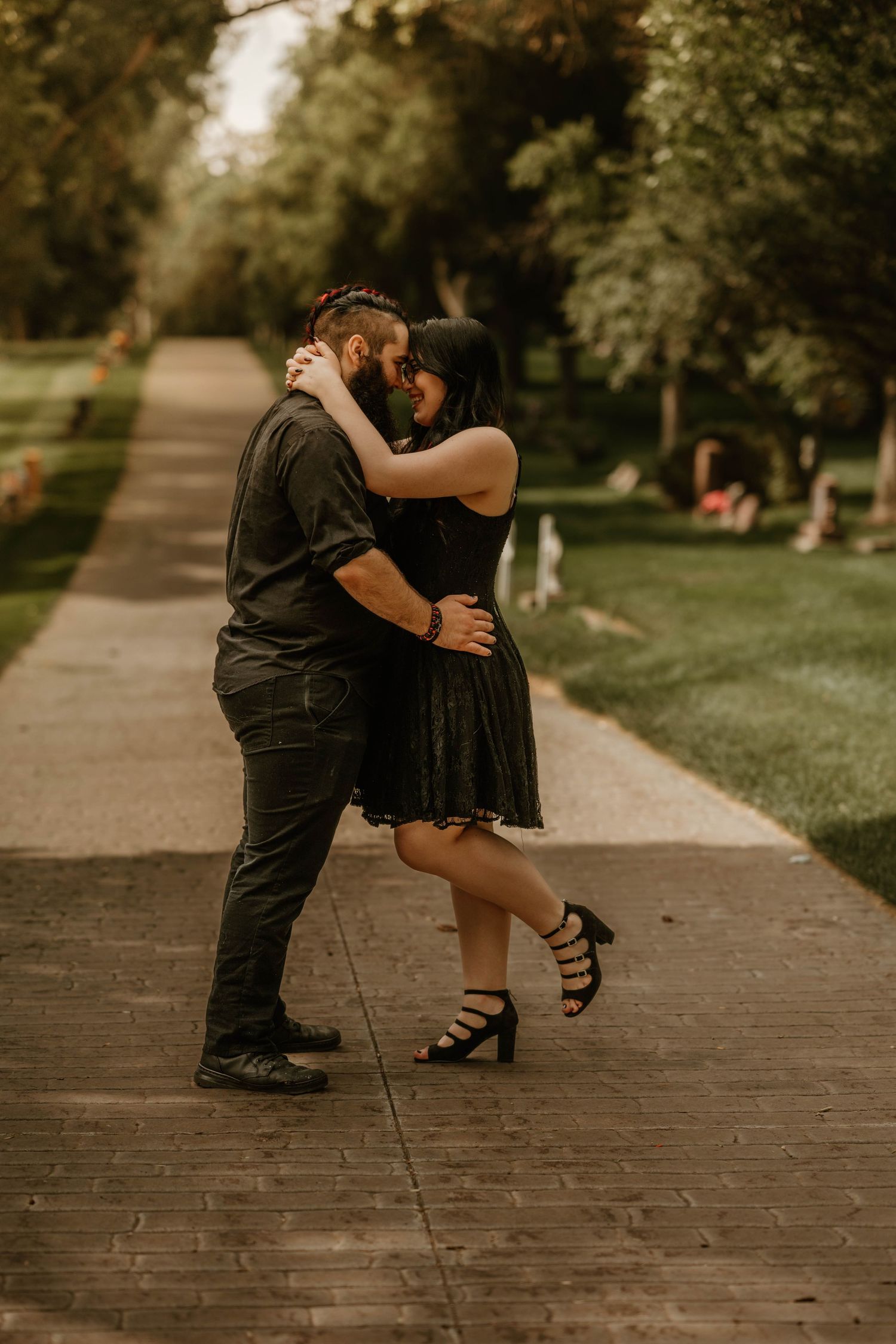 Dancing couple in black outfits performing romantic dance moves on a brick pathway in a park.