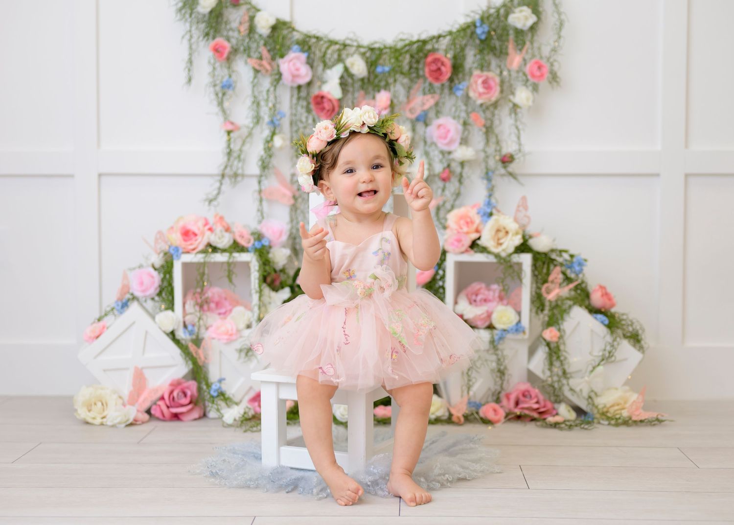 A festive pink and floral birthday setup with a tutu-wearing subject seated on a small white chair against a decorated wall.