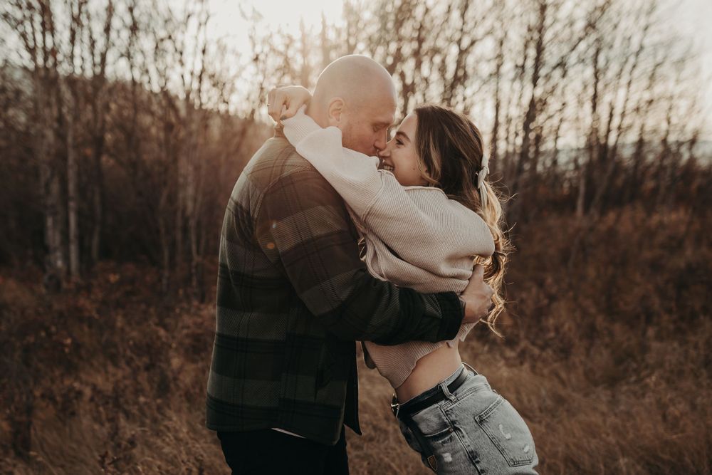 A romantic couple embraces in a natural setting during golden hour with trees in the background.