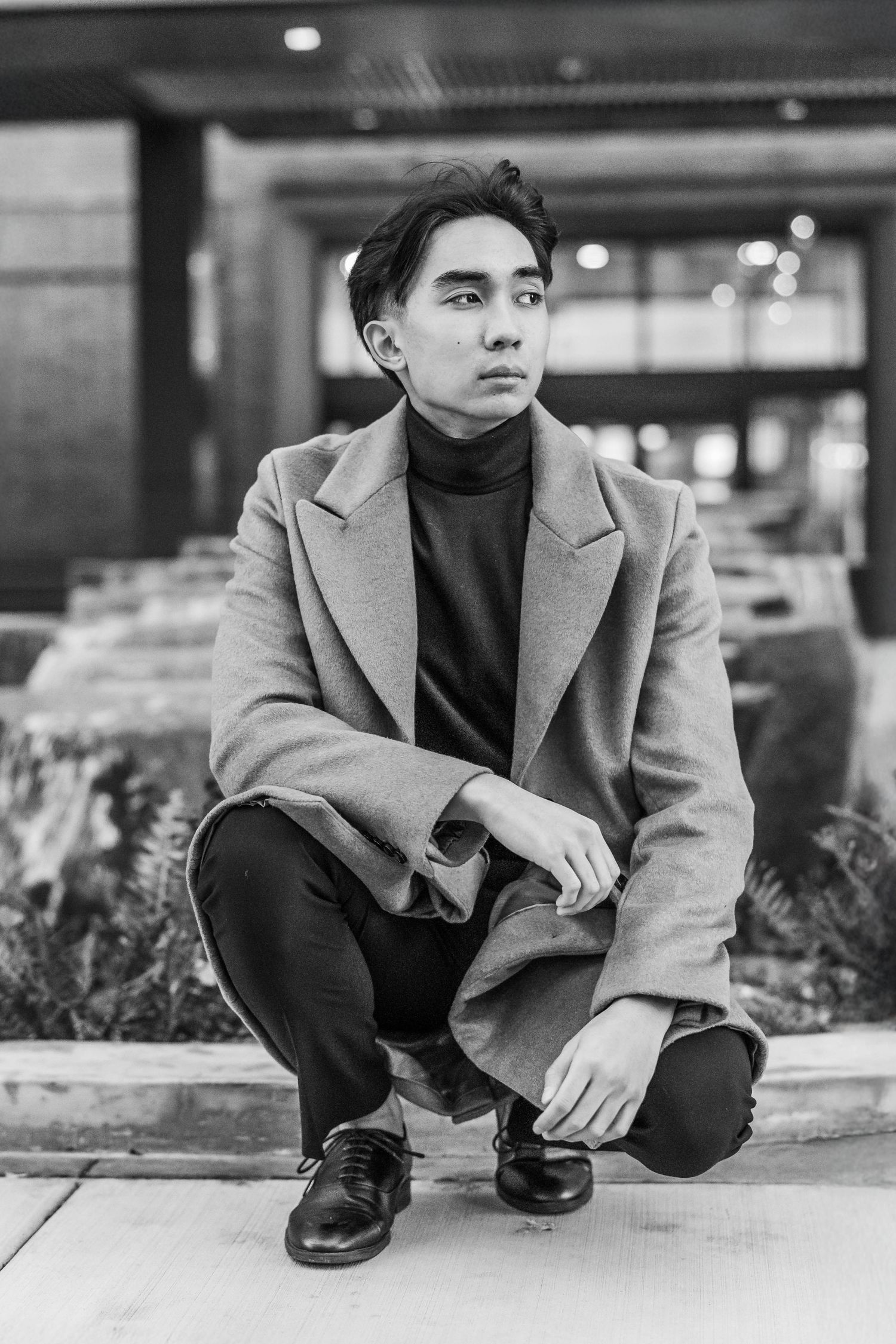 Young Asian man in camel coat crouching outdoors in black and white urban portrait photo.