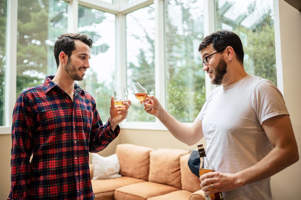 Two men toasting with whiskey glasses and a beer bottle in a bright living room, smiling and celebrating together.