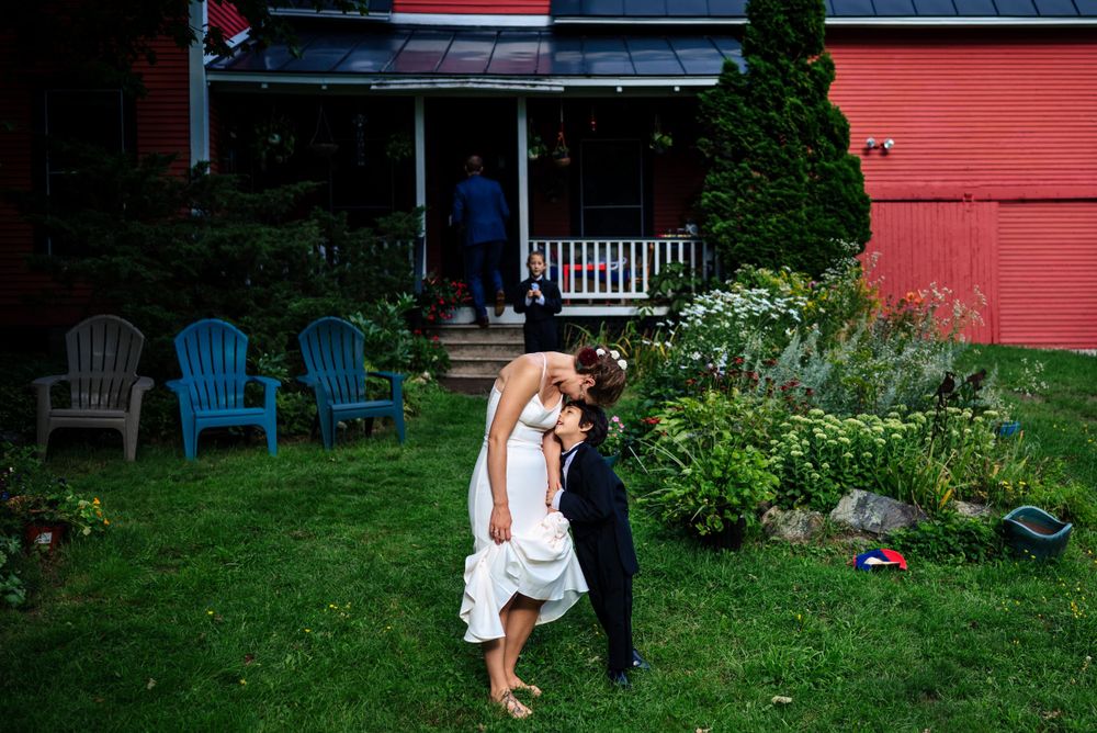 Bride and groom share romantic dip kiss on green lawn beside red barn farmhouse.