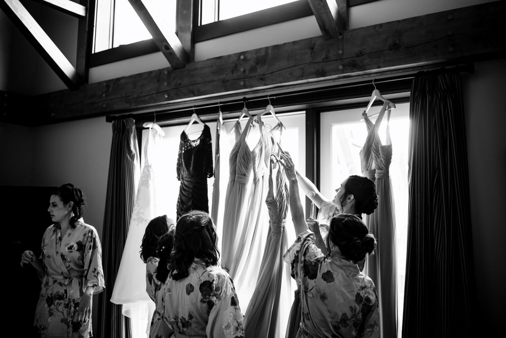 Bridesmaids in floral robes admire hanging dresses backlit by bright windows, black and white.