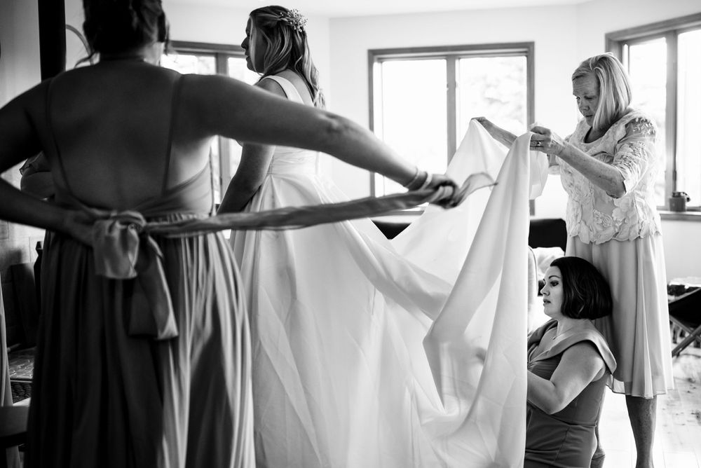 Bridesmaids help bride into wedding dress and veil in bright dressing room, black and white.