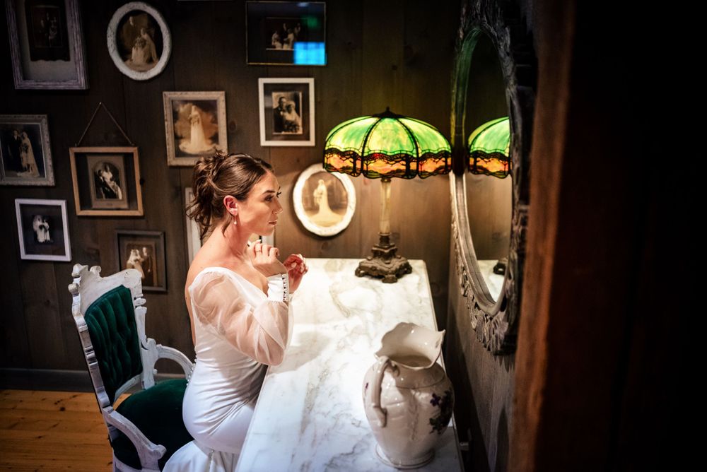 Bride in white gown applies makeup at vintage vanity table with green stained glass lamp.