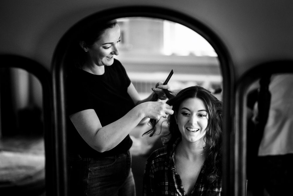 Smiling bride gets hair styled by hairdresser captured in black and white mirror reflection.