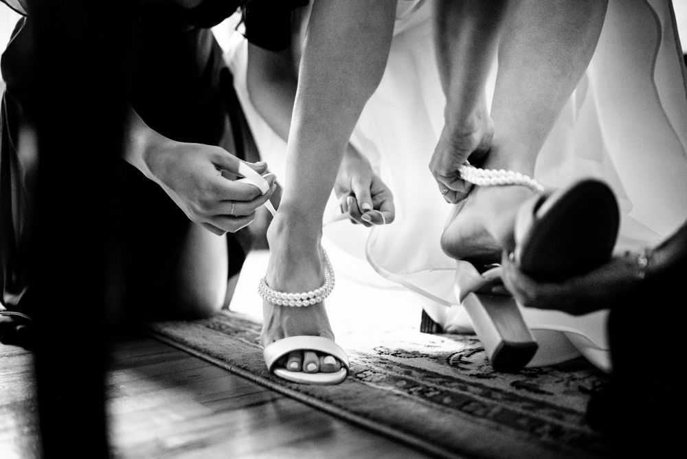 Black and white photo of bride getting wedding shoes fastened by assistant.