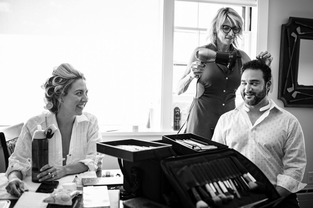 Bride laughs with groom and makeup artist during fun wedding morning preparations, black and white.