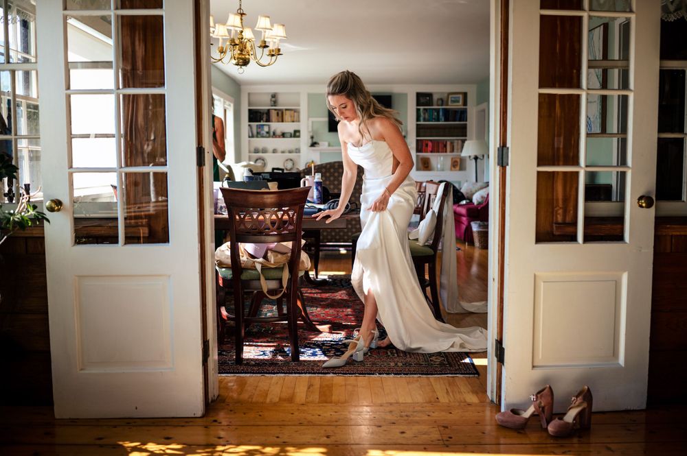 Bride in white gown standing in sunlit room with French doors.
