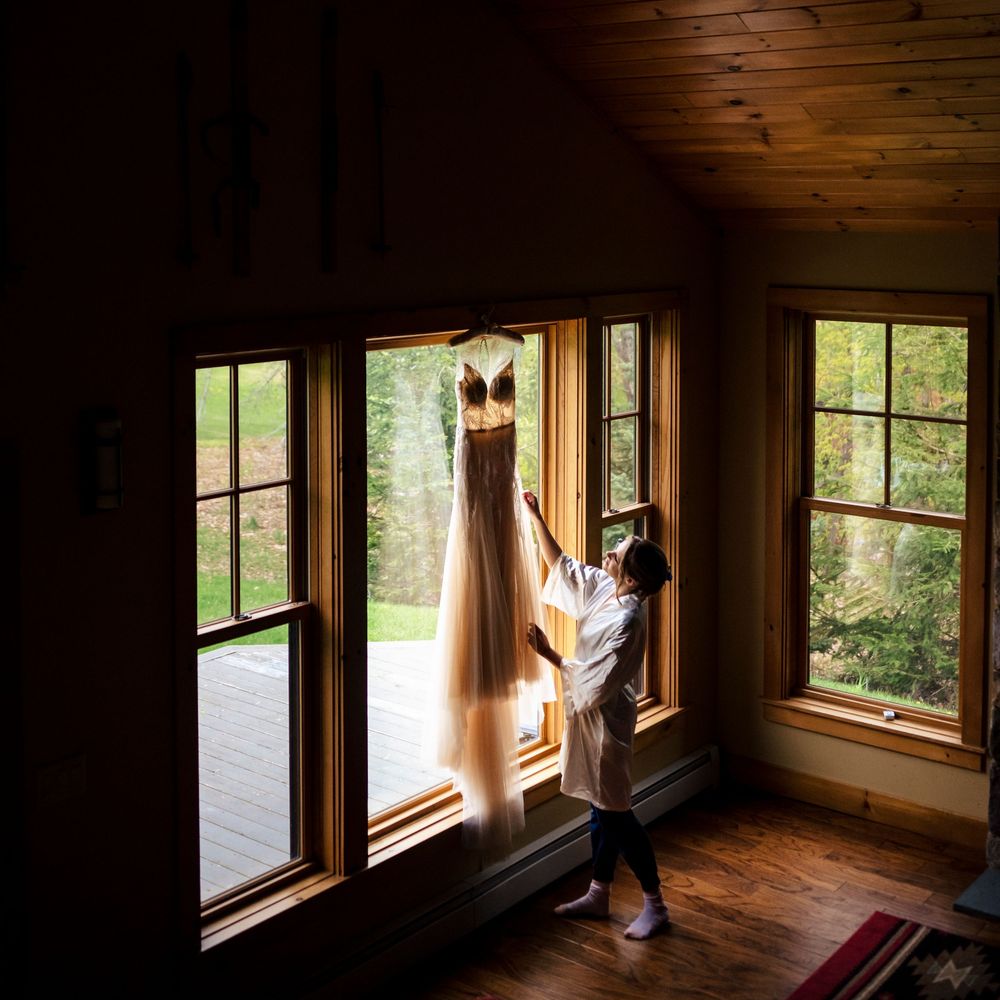 Bride adjusting her wedding gown hanging by sunlit cabin windows with wooden floors and rustic decor.
