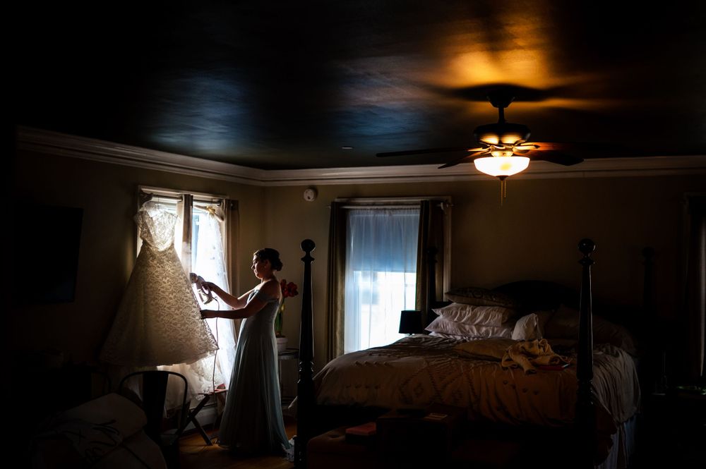 Bride examining wedding dress hanging by window in dimly lit bedroom.