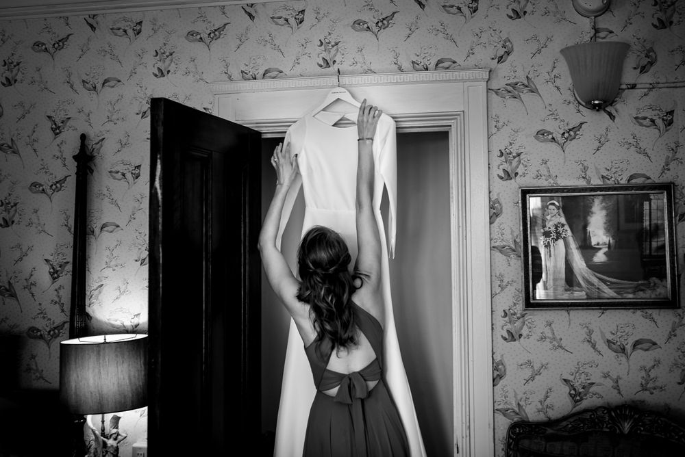 Bride reaches for wedding dress on hanger in vintage wallpapered room, black and white photo.