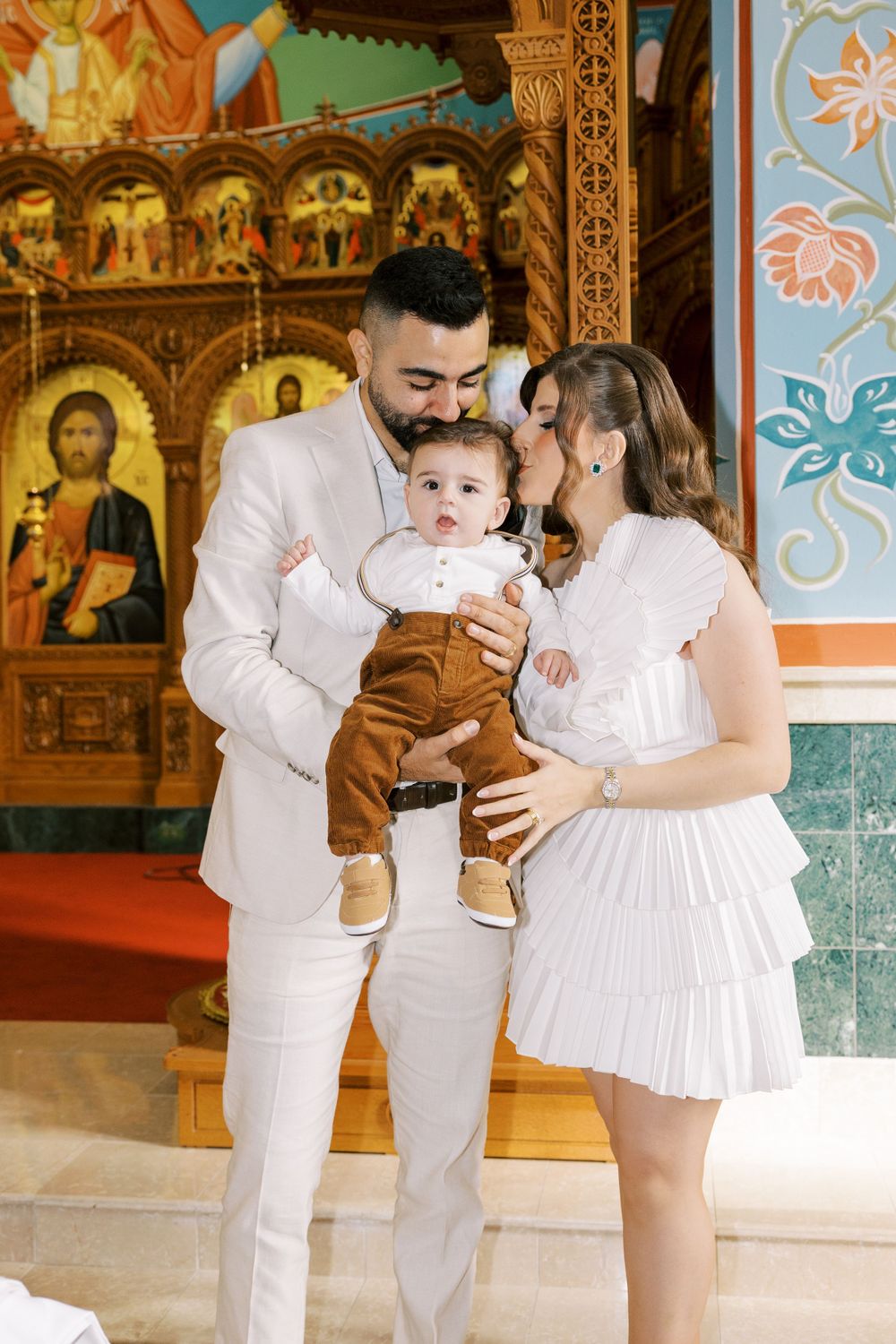 Parents kissing their smiling baby boy during Orthodox baptism ceremony inside decorated church.