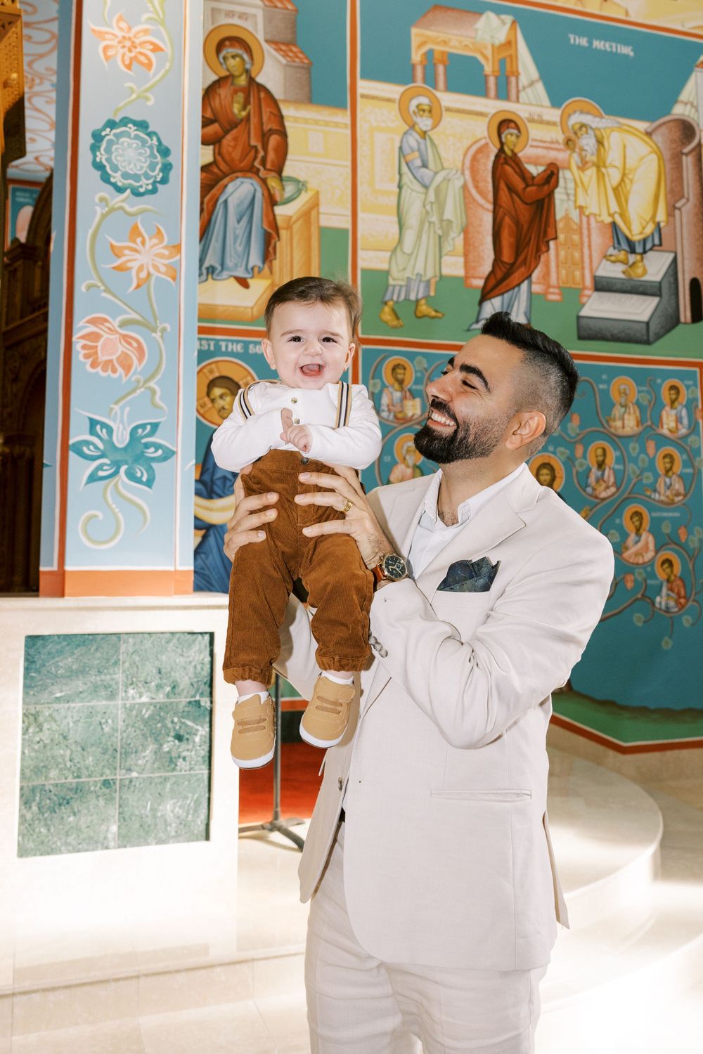Father in white suit lifting laughing baby boy in front of colorful Orthodox church murals.