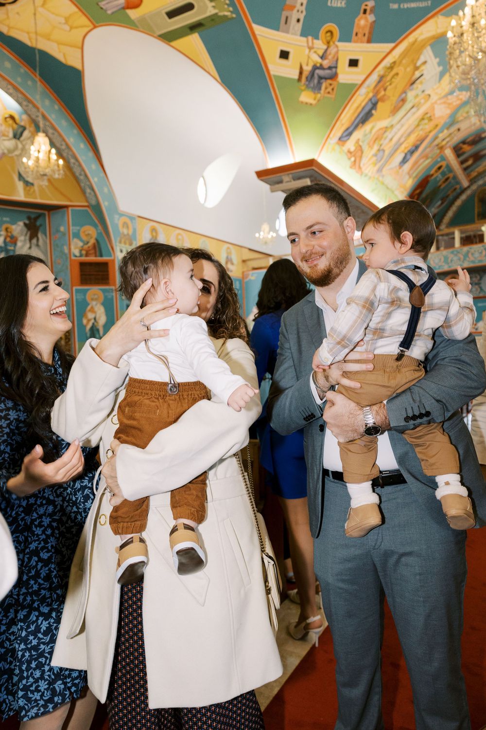 Guests celebrate at Orthodox church baptism holding two babies surrounded by colorful Byzantine frescoes.