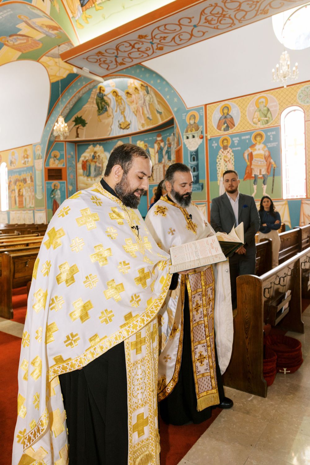 Two Orthodox priests in white and gold vestments read from scripture during baptism ceremony.