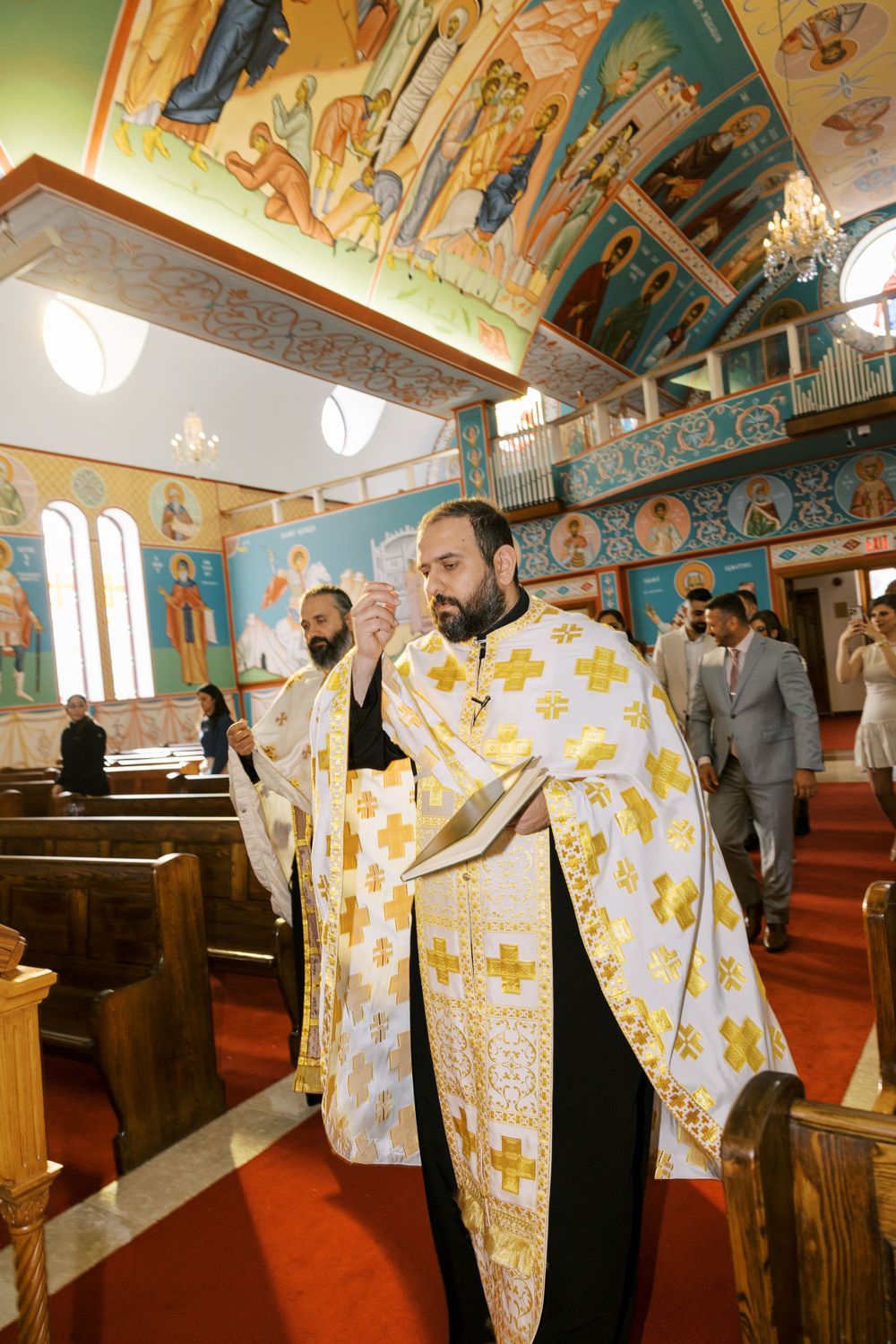 Orthodox priest in white and gold vestments walks down aisle of colorfully decorated Byzantine church.
