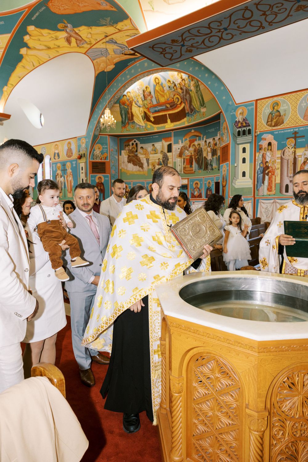 Orthodox priest holds scripture book over baptismal font during christening ceremony with family gathered.
