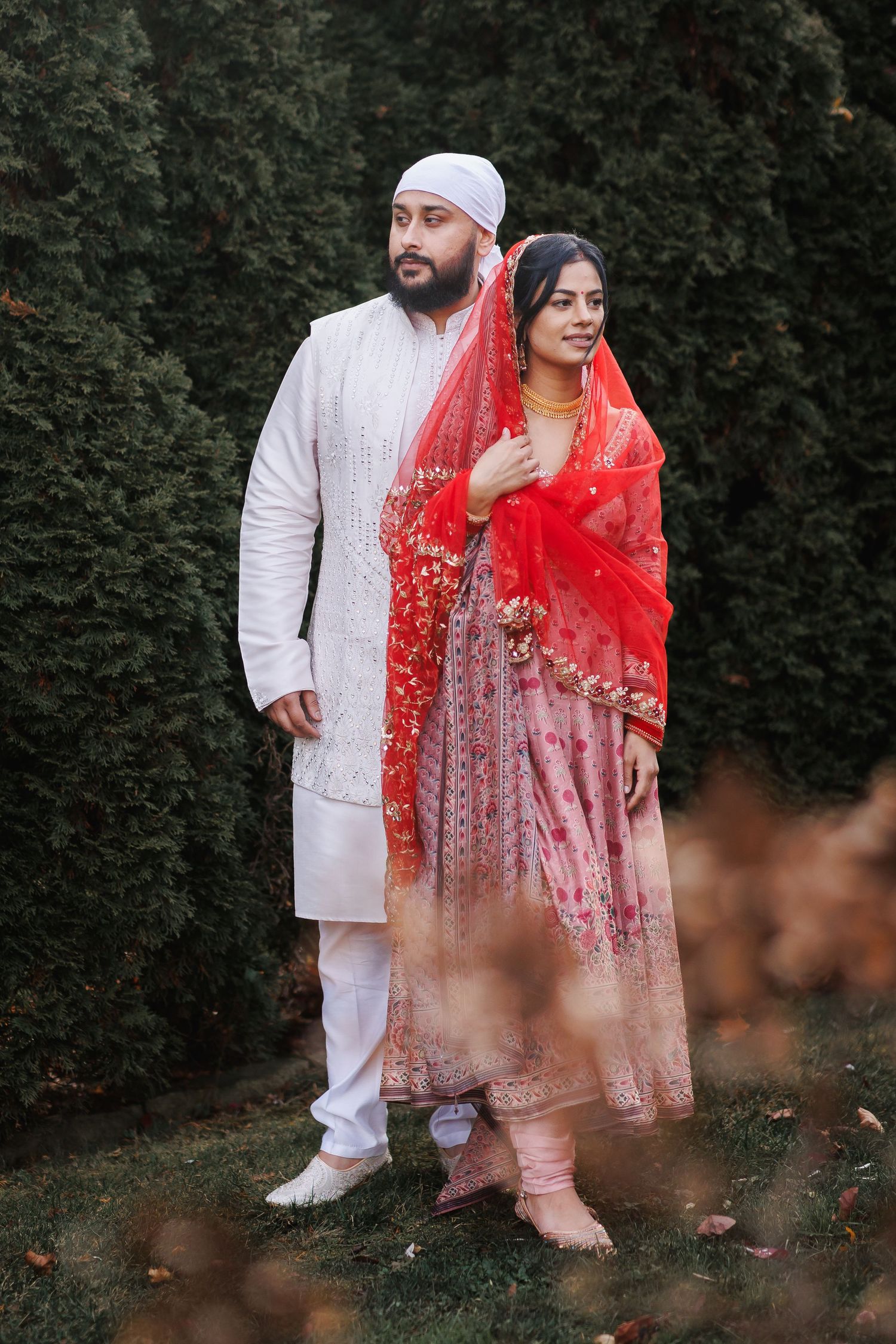 A couple in traditional South Asian wedding attire poses against a hedge backdrop.