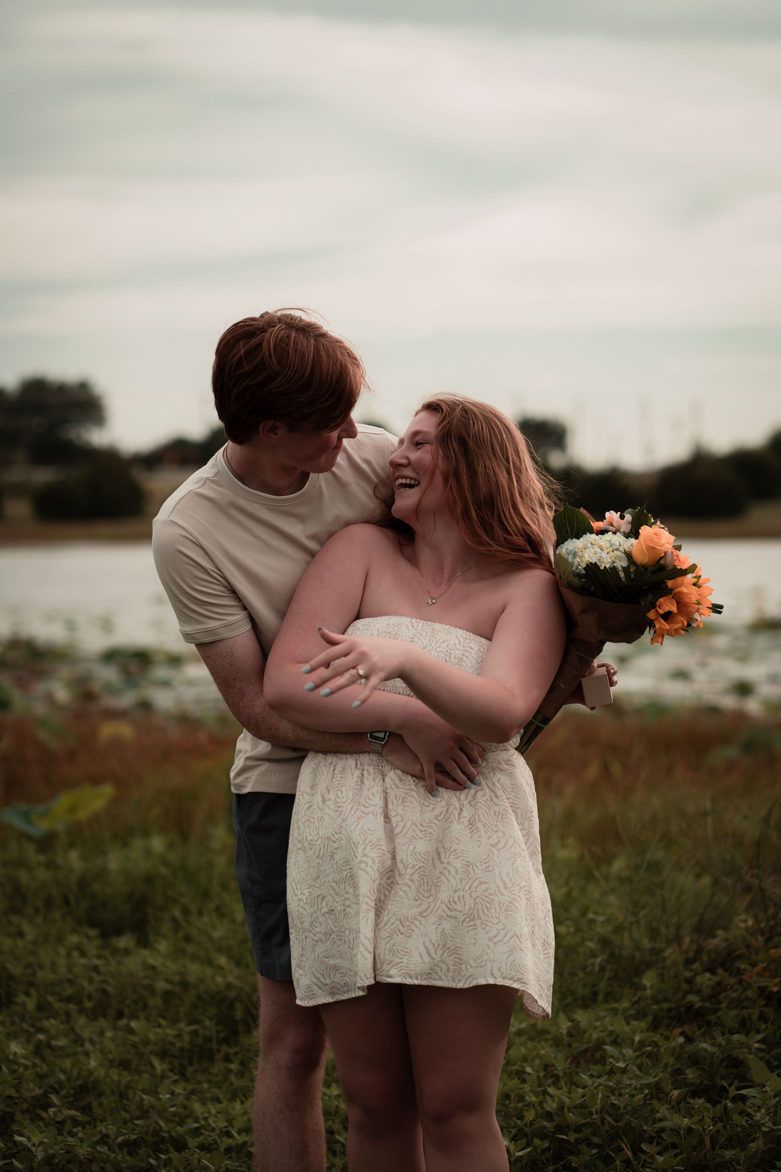A couple embraces in a field at sunset with orange and white flowers in their hands.
