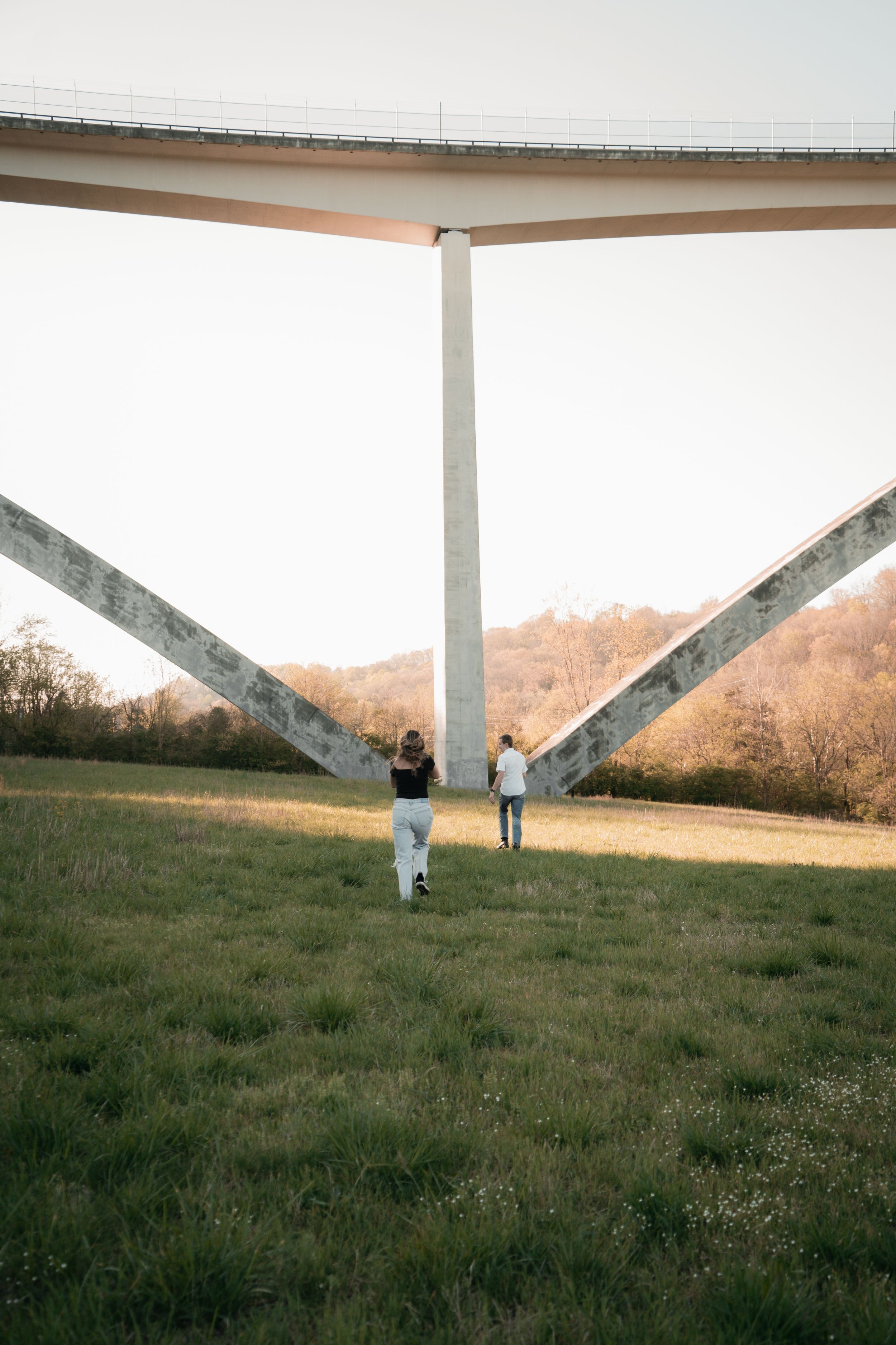 Two people walk beneath a large concrete highway overpass structure in a grassy field at sunset.